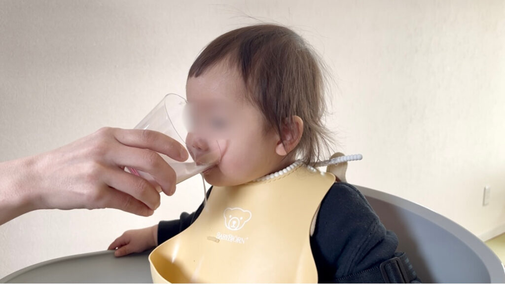 大人のガラス素材のコップで牛乳を飲まされている子供の様子。決して自分ではグラスを持たず、ママがグラスを持っています。よくみると少しこぼしています。